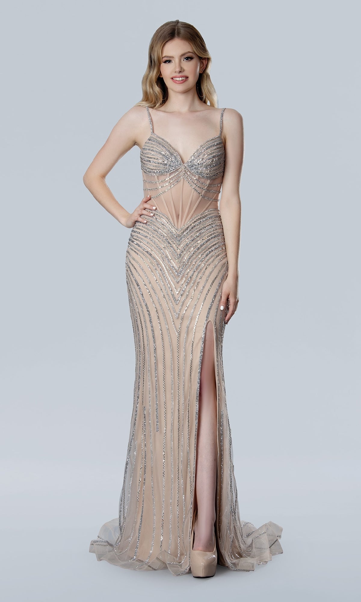 A woman stands against a plain light background wearing the Stella Couture 25198, a floor-length, nude form-fitting prom dress with silver sequin details, thin straps, and a high slit.